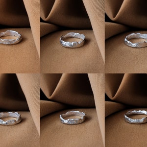 May include: A silver ring with a textured, organic design. The ring is displayed on a brown fabric background.