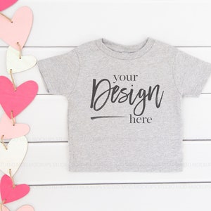 Könnte beinhalten: Ein graues T-Shirt mit kurzen Ärmeln mit dem Text "your design here" auf einem weißen Holzgrund mit einer Girlande aus rosa und weißen Herzen.