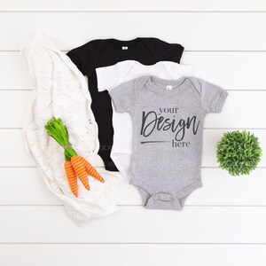 May include: Three baby onesies, one black, one white and one grey. The grey onesie has the text "Your Design Here" printed on it. The onesies are arranged on a white wooden surface with a white knitted blanket, carrots and a small green plant.