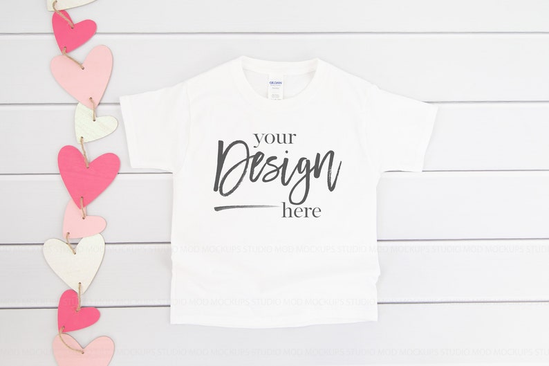May include: White t-shirt with the text "your design here" on a white wood background with a pink and white heart garland.