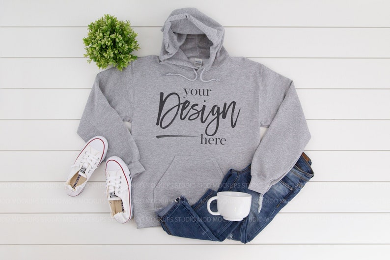 May include: A gray hooded sweatshirt with a pocket on the front. The sweatshirt has the text "your design here" printed on the front. The sweatshirt is laid out on a white wooden surface with a pair of white sneakers, blue jeans, and a white mug.