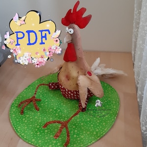 Può includere: Un pollo marrone e beige con cresta e bargigli rossi, seduto su un tappetino rotondo verde con pois bianchi. Il pollo ha una coda di piume bianche e zampe marroni. Il tappetino ha il testo "lilhodimaisl@gmail.com" e "Chiarra Mereschko" in nero.