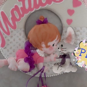 Puede incluir: Una corona de fieltro con el nombre "Matilda" en letras rosas. Una muñeca vestida de rosa y morado con cabello castaño y un conejito blanco con orejas grises están en el centro de la corona. La corona está decorada con cuentas blancas y una cinta rosa y morada.