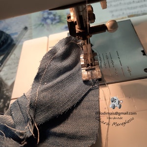 May include: A close-up of a sewing machine needle going through a piece of blue denim fabric. The machine has a white plastic foot and a white plastic knob. The fabric is folded over and the needle is going through the fold. The text "ilfilodimmais@gmail.com handmade Chiara Meneghesso" is visible in the lower right corner of the image.