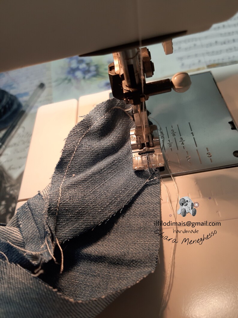 May include: A close-up of a sewing machine needle going through a piece of blue denim fabric. The machine has a white plastic foot and a white plastic knob. The fabric is folded over and the needle is going through the fold. The text "ilfilodimmais@gmail.com handmade Chiara Meneghesso" is visible in the lower right corner of the image.
