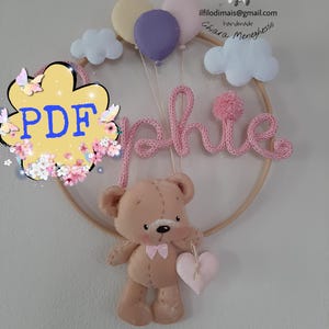 Puede incluir: Decoración de guardería hecha a mano con un osito de peluche sosteniendo un corazón rosa, un aro de madera y el nombre "Sophie" en hilo rosa. El diseño incluye nubes de fieltro y globos en colores pastel.