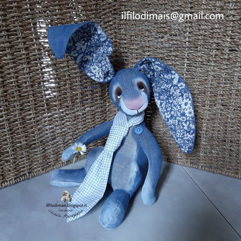 May include: A blue denim bunny rabbit with a white and blue checkered scarf, holding a white daisy. The bunny's ears are lined with a white floral pattern. The bunny is sitting on a light gray surface.