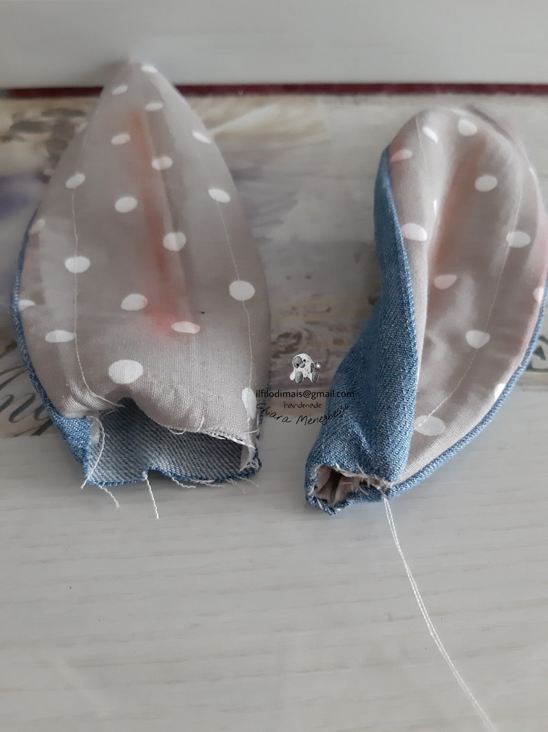 May include: Two gray fabric bunny ears with white polka dots and blue denim trim. The ears are sewn together at the base and have a white thread attached.