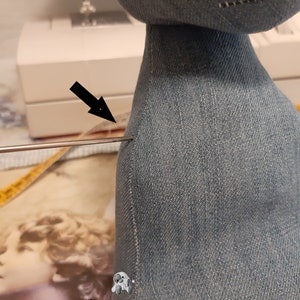 May include: Close-up of a denim fabric doll with a seam along the back. A needle is inserted through the fabric, indicating a sewing project.