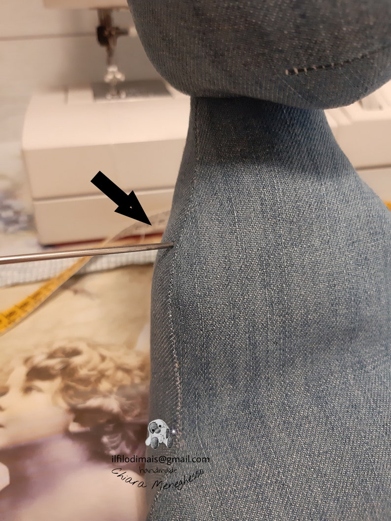 May include: Close-up of a denim fabric doll with a seam along the back. A needle is inserted through the fabric, indicating a sewing project.