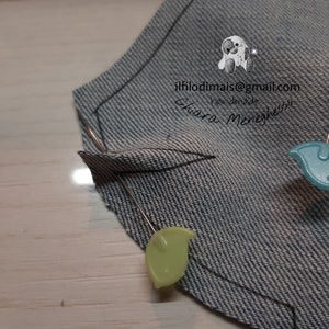 May include: A close-up of a denim fabric piece with a curved line drawn on it. Two pins with green and blue plastic heads are securing the fabric.