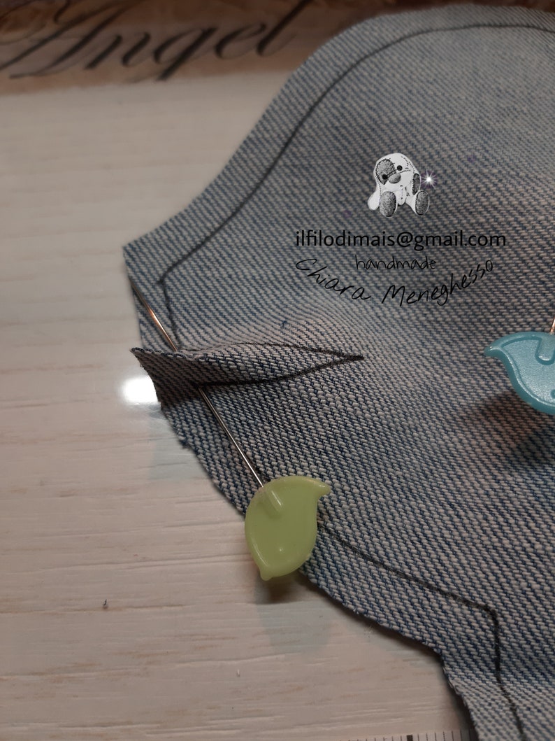 May include: A close-up of a denim fabric piece with a curved line drawn on it. Two pins with green and blue plastic heads are securing the fabric.