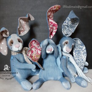 May include: Three handmade denim bunny rabbits with floral fabric accents. The bunnies are sitting on a light brown surface. The bunnies have white polka dot ears, blue and white floral ears, and white and blue floral ears.