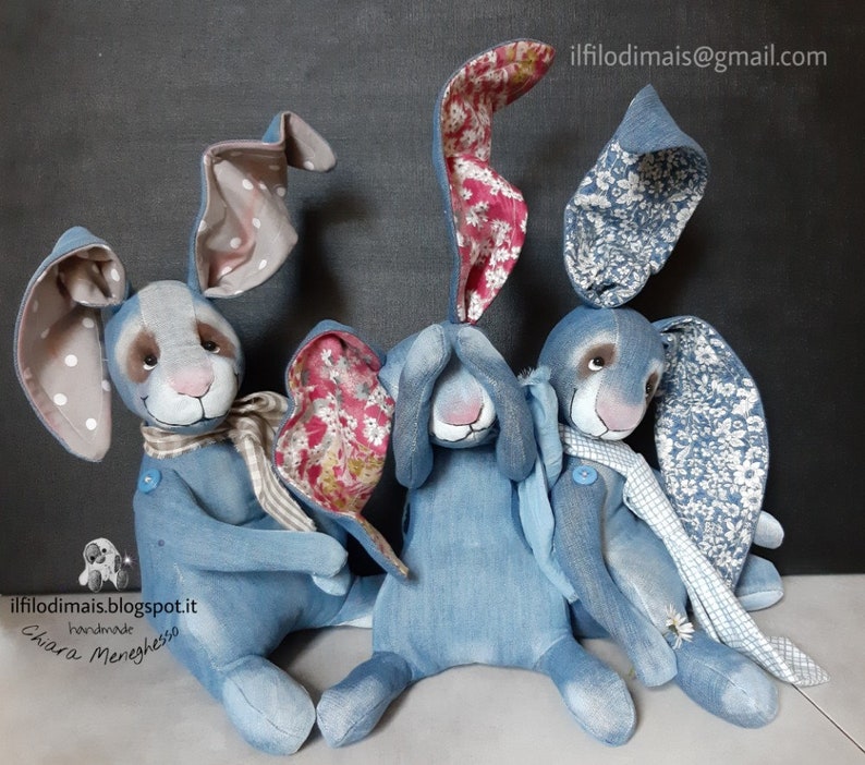 May include: Three handmade denim bunny rabbits with floral fabric accents. The bunnies are sitting on a light brown surface. The bunnies have white polka dot ears, blue and white floral ears, and white and blue floral ears.