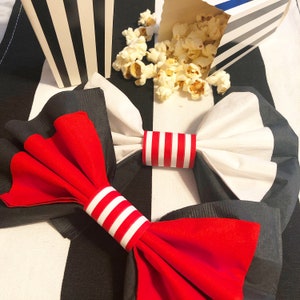 May include: A festive arrangement featuring red and white bow-tie napkins with striped napkin rings. A box of popcorn and a black and white striped container are also present, all set against a black and white checkered background. The theme is a fun, celebratory event.