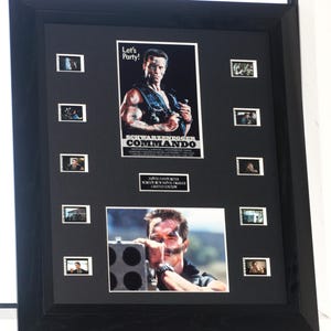 May include: Framed movie poster and film stills from the movie Commando, featuring Arnold Schwarzenegger. The poster has the text "Let's Party!" and "Commando". The frame is black.