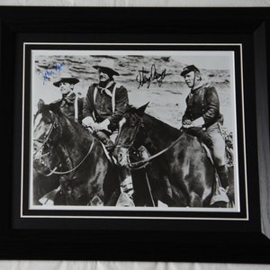 Peut inclure: Photographie en noir et blanc de trois hommes à cheval. Les hommes portent des vêtements d'époque et des chapeaux. La photo est signée par les trois hommes, "John Wayne", "Harry Morgan" et "Strother Martin".