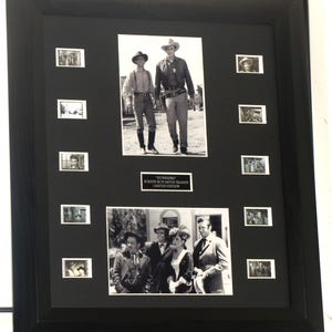 May include: A black framed display featuring two black and white photographs from the television show Gunsmoke, surrounded by eight small film cells. A central plaque reads: GUNSMOKE SCREEN RUN MOVIE FRAMES LIMITED EDITION. The display is mounted on a black mat.