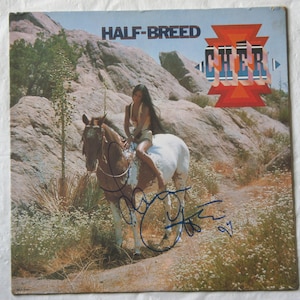 Könnte beinhalten: Albumcover für Cher's "Half-Breed" mit Cher auf einem Pferd in einer Wüstenlandschaft. Der Albumtitel und der Künstlername sind in einer stilisierten Schrift dargestellt. Das Cover ist mit blauem Stift signiert.