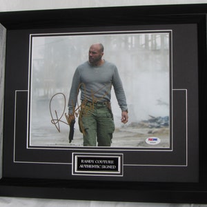 Peut inclure: Une photo en noir et blanc de Randy Couture, un combattant d'arts martiaux mixtes, dans une scène de film. Il porte une chemise grise, un pantalon vert et un holster d'arme à feu. Sa signature est en encre dorée sur la photo. La photo est encadrée en noir et comporte un autocollant d'authentification PSA/DNA.