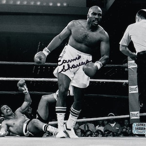 Pode incluir: Fotografia em preto e branco de um boxeador em um ringue de boxe. O boxeador está vestindo shorts brancos e está lançando um soco. O outro boxeador está no chão. A foto é assinada por "Earnie Shavers".