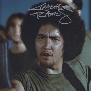 May include: A vintage photograph featuring a man with a large afro and a serious expression. The man is wearing a green shirt and appears to be holding an object. The image is signed in white ink.
