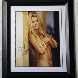 May include: Framed photograph featuring a woman with long blonde hair, wearing minimal clothing. The image is signed in black ink. The photograph is set within a white mat and a black frame.