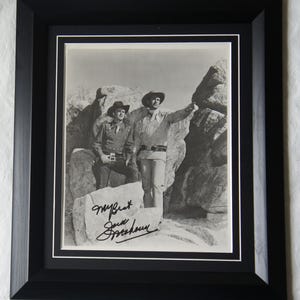 Op de afbeelding: Zwart-wit ingelijste foto met twee mannen in cowboykleding, die buiten poseren. De foto is gesigneerd op een rots op de voorgrond. De lijst is zwart met een witte passe-partout.
