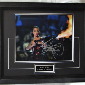 Puede incluir: Una foto enmarcada en negro de Ruby Rose sosteniendo un lanzallamas, con su firma en tinta negra. La foto está enmarcada y tiene una placa plateada que dice "Ruby Rose Authentic Signed".