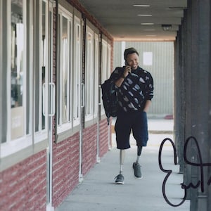 Könnte beinhalten: Eine Person in einem schwarz-weiß gemusterten Sweatshirt, marineblauen Shorts und grauen Turnschuhen geht einen Flur entlang. Sie trägt einen Rucksack und telefoniert. Die Person hat ein Beinprothese. Das Bild ist signiert.