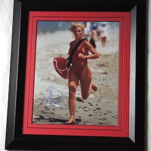 May include: Framed photograph of a woman in a red swimsuit running on a beach, carrying a rescue buoy. The photo is in a black frame with a red matting, and includes an autograph.