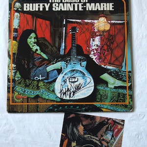 Könnte beinhalten: Vintage Vinyl-Schallplattenhülle für "The Best Of Buffy Sainte-Marie". Das Cover zeigt eine Frau mit langen dunklen Haaren, eine Gitarre und ein Autogramm. Das Albumcover hat einen orangefarbenen Rand und der Hintergrund ist rot.