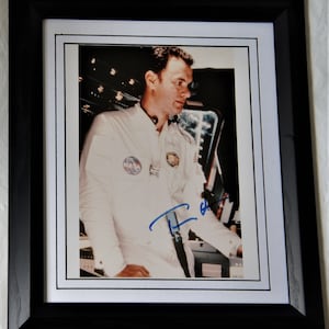 May include: Framed photograph of an astronaut in a white spacesuit, featuring the NASA logo. The image is signed in blue ink. The photograph is set within a white mat and a black wooden frame.