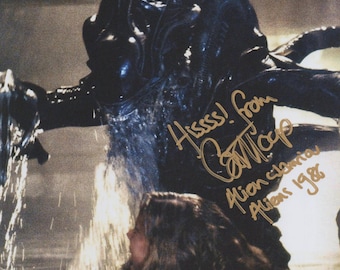 May include: A dark, menacing creature from the movie "Aliens" with a signature and the text "Hiss!" and "Alien warrior Aliens 1986". The creature is black and has a textured, alien-like appearance. A person is in the foreground.