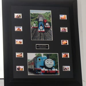 Puede incluir: Un marco negro que presenta dos escenas de la película Thomas y sus amigos, ocho pequeñas celdas de película y una placa central que dice MOVIE FAVOURITES SCREEN RUN MOVIE FRAMED LIMITED EDITION. La imagen superior muestra tres trenes.