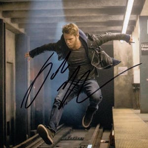May include: A man wearing a black leather jacket and jeans jumps in mid-air with his arms outstretched. The image is signed with the name "John" in black ink.
