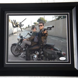 May include: Framed photograph featuring two people on a motorcycle, one holding a shotgun. The image is signed in black ink. The photograph is in a black frame with a white mat.