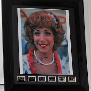 May include: Framed photograph of a smiling woman with curly red hair, wearing a white beaded necklace and a red lei. The photo is signed in blue ink. The black frame includes a row of small film strip images.