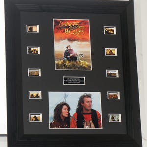 May include: A framed "Dances with Wolves" movie display. The black frame contains a central movie poster and several small film stills. The poster shows a man and a dog on a grassy hill with a sunset. Text reads "Dances with Wolves".