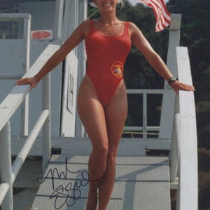 May include: A woman in a red lifeguard swimsuit stands on a wooden structure, arms outstretched. The swimsuit features a circular emblem. An American flag waves in the background. The image is signed.