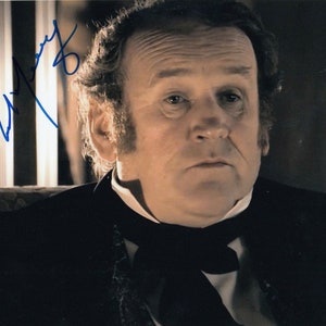 May include: A close-up portrait of a man with a serious expression. He is wearing a black suit jacket with a white shirt and a black bow tie. The image is signed "Anthony Hopkins".