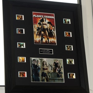 Op de afbeelding: Een zwart ingelijst filmposter en filmceldisplay met de film "Planet Terror". De poster toont een vrouw in een rode bikini met een pistool. De filmcellen tonen scènes uit de film.
