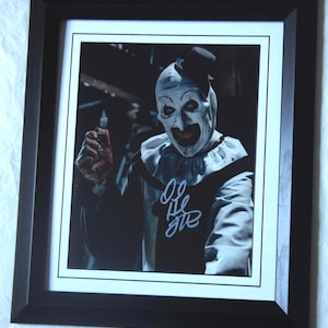 Op de afbeelding: Een ingelijste foto van een clown met een wit gezicht en rode lippen, die een zwart-wit kostuum draagt. De clown houdt een bloedig mes vast en kijkt naar de camera. De foto is gesigneerd met "Art the Clown".