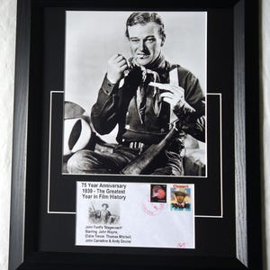 Peut inclure: Photographie encadrée en noir et blanc de John Wayne, avec un cadre noir et un passe-partout blanc. L'image comprend une enveloppe commémorative avec des timbres et le texte : "75e anniversaire 1939 - La plus grande année de l'histoire du cinéma."