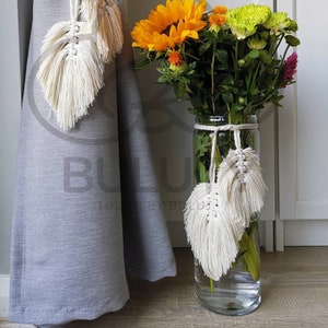 Peut inclure: Deux glands de macramé en forme de feuille blanche attachés à un rideau gris et un vase en verre transparent avec un bouquet de tournesols jaunes, de fleurs vertes et de fleurs orange. Le vase est également attaché avec un gland de macramé en forme de feuille blanche.
