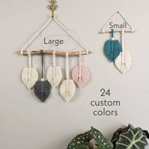 May include: Two macrame wall hangings with a wooden branch, one labeled "Large" and the other "Small". The large wall hanging has five feather-shaped tassels in white, gray, and pink. The small wall hanging has two feather-shaped tassels in teal and white. The text "24 custom colors" is below the wall hangings.