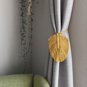 Peut inclure: Un lien de rideau en macramé en forme de feuille jaune moutarde suspendu à un rideau rayé gris. Le lien est fait de fil épais et a une frange en bas.