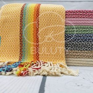 May include: A stack of colorful Turkish towels with a rainbow stripe design. The towels are made of cotton and have a fringe border. The towels are folded and stacked on a white wooden surface.