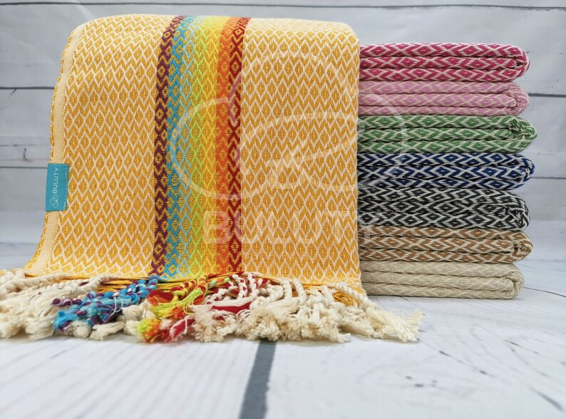 May include: A stack of colorful Turkish towels with a rainbow stripe design. The towels are made of cotton and have a fringe border. The towels are folded and stacked on a white wooden surface.