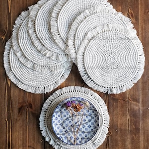 May include: Set of eight white crocheted placemats with fringe trim. The placemats are arranged in a circle with one placemat in the center featuring a blue and white patterned plate with dried flowers.